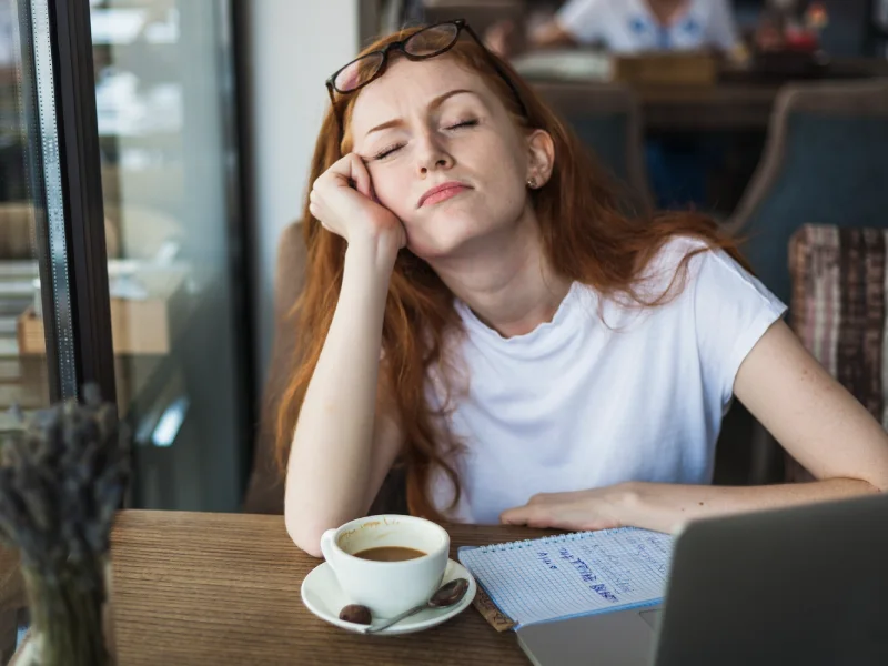 uma mulher cansada com os olhos fechados e uma chávena de café pousada numa mesa
